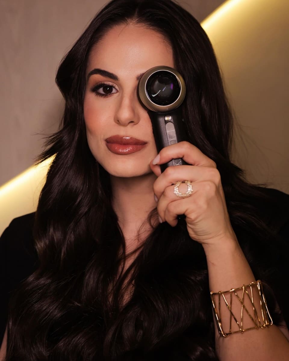 Dra. Caroline Del Colle em retrato de apresentação, segurando um instrumento de exame junto ao rosto.